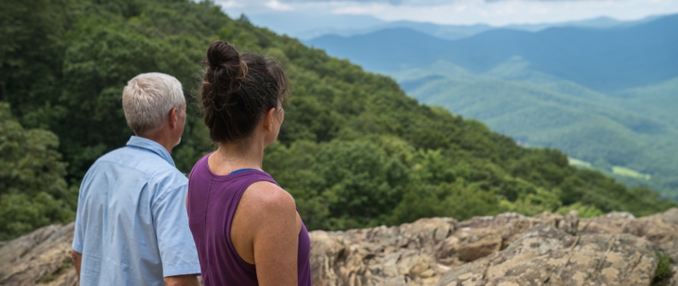 A male and female couple overlooks mountains and hills covered in greenery.