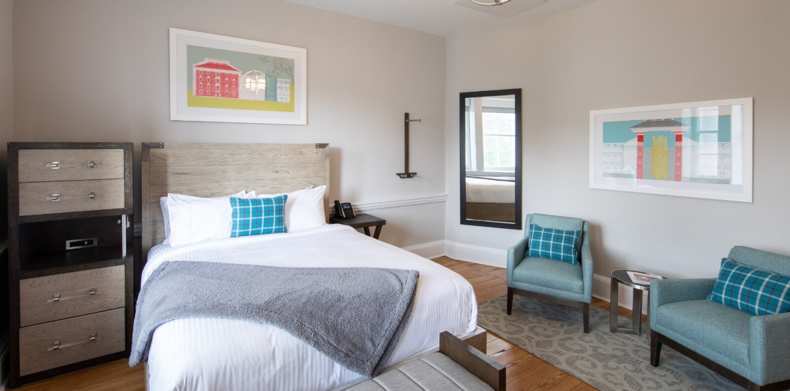 A guest room at the Blackburn Inn in Staunton Virginia is decorated with light wood furniture, blue chairs and white linens. Colorful paintings and a full size mirror hang on the wall. 